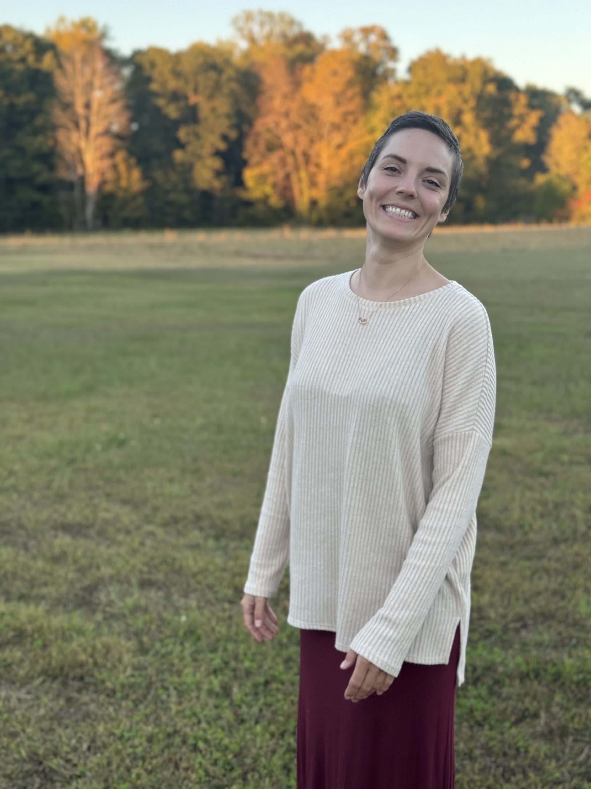 A smiling woman in a light sweater standing outdoors in a sunlit field with trees in the background.