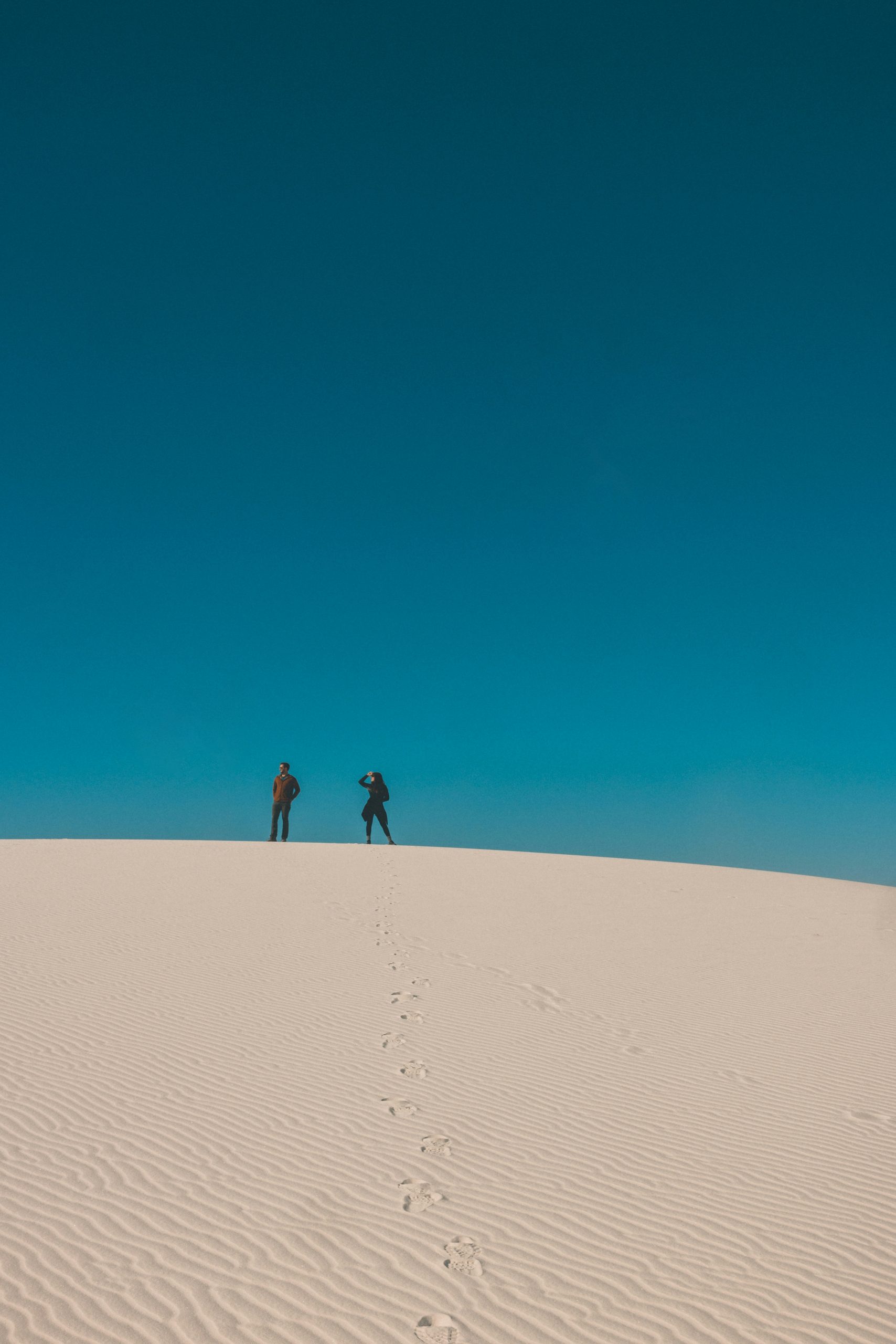 Two figures walking across a vast sand dune under a clear blue sky.