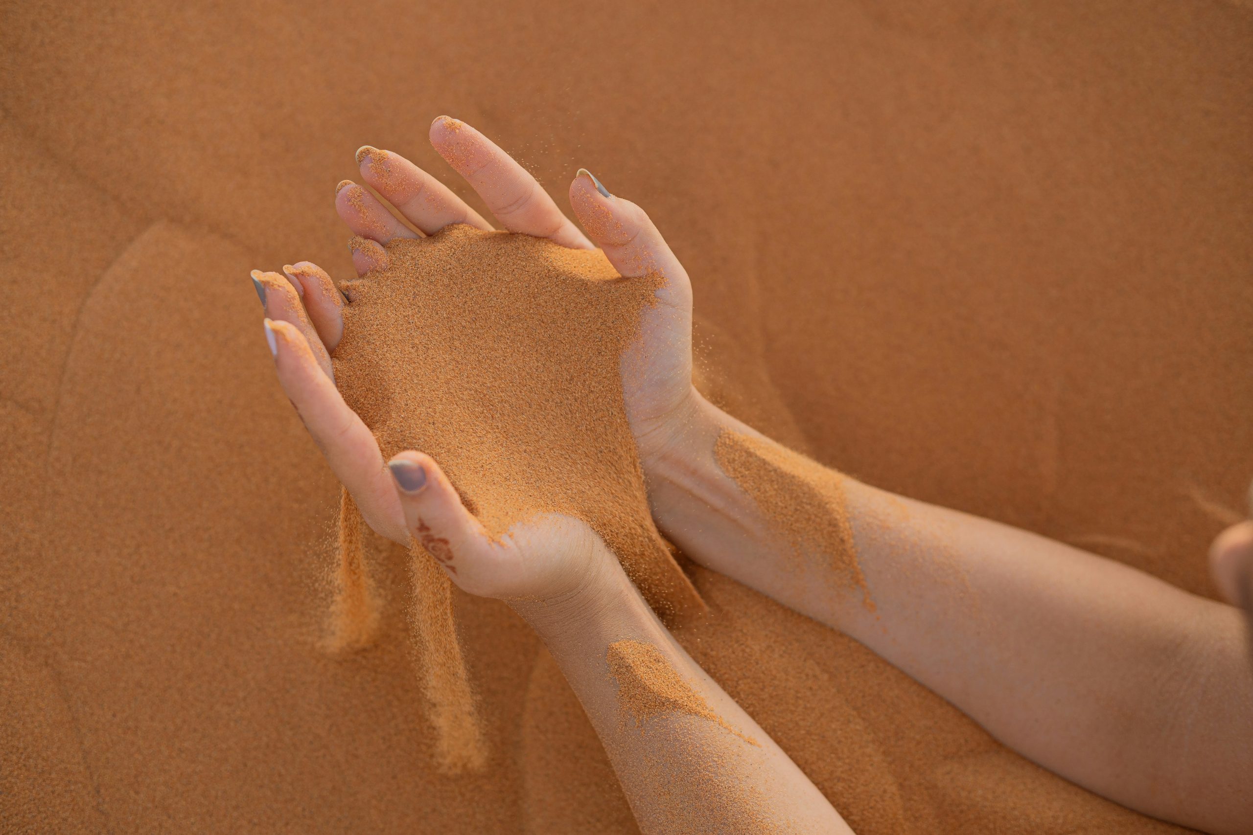 Woman with medium length nails and nail polishes lets light orange sand spill from her hands with nothing but sand in the background