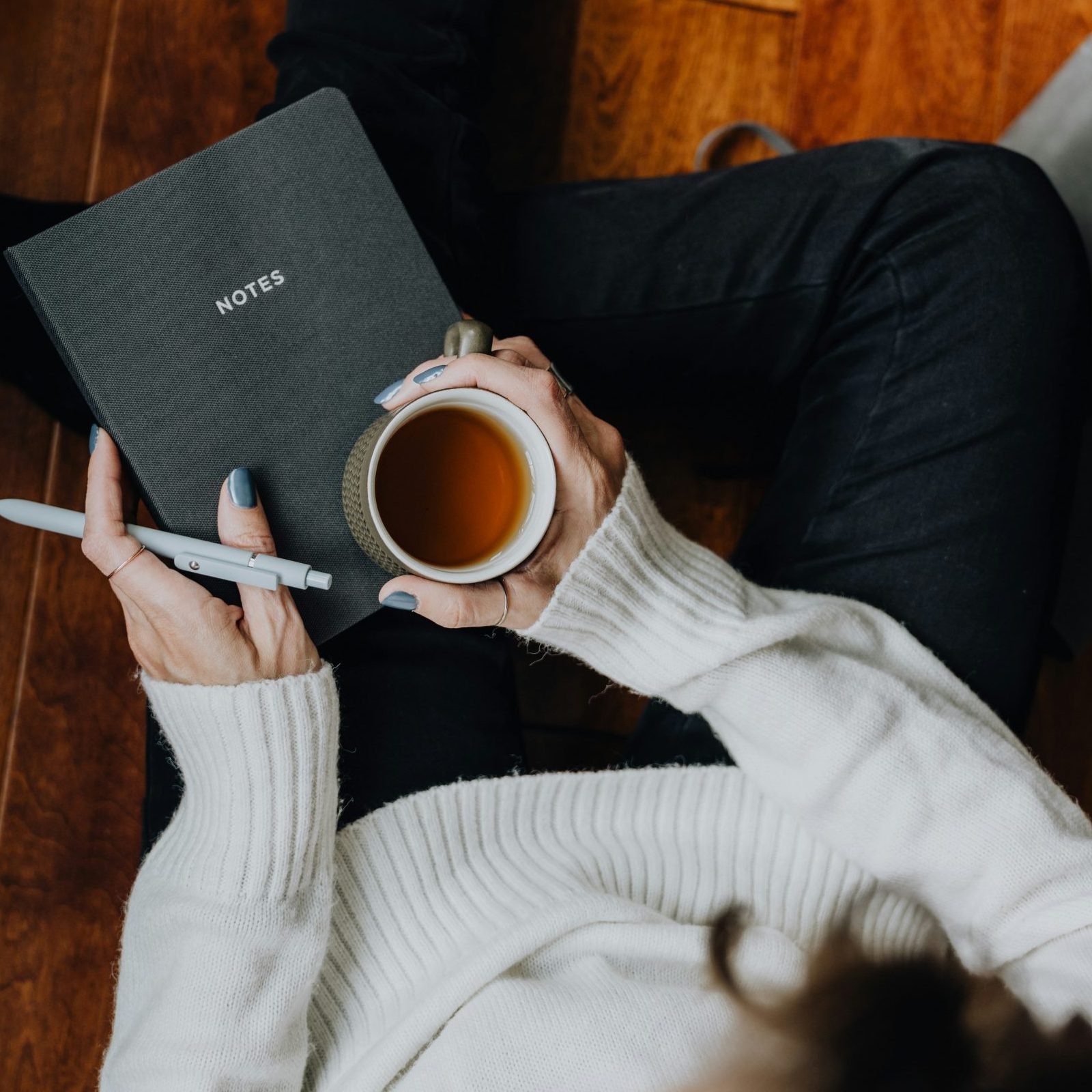 A cozy overhead view of a woman in a cream sweater holding a cup of tea in one hand and an open journal in the other.