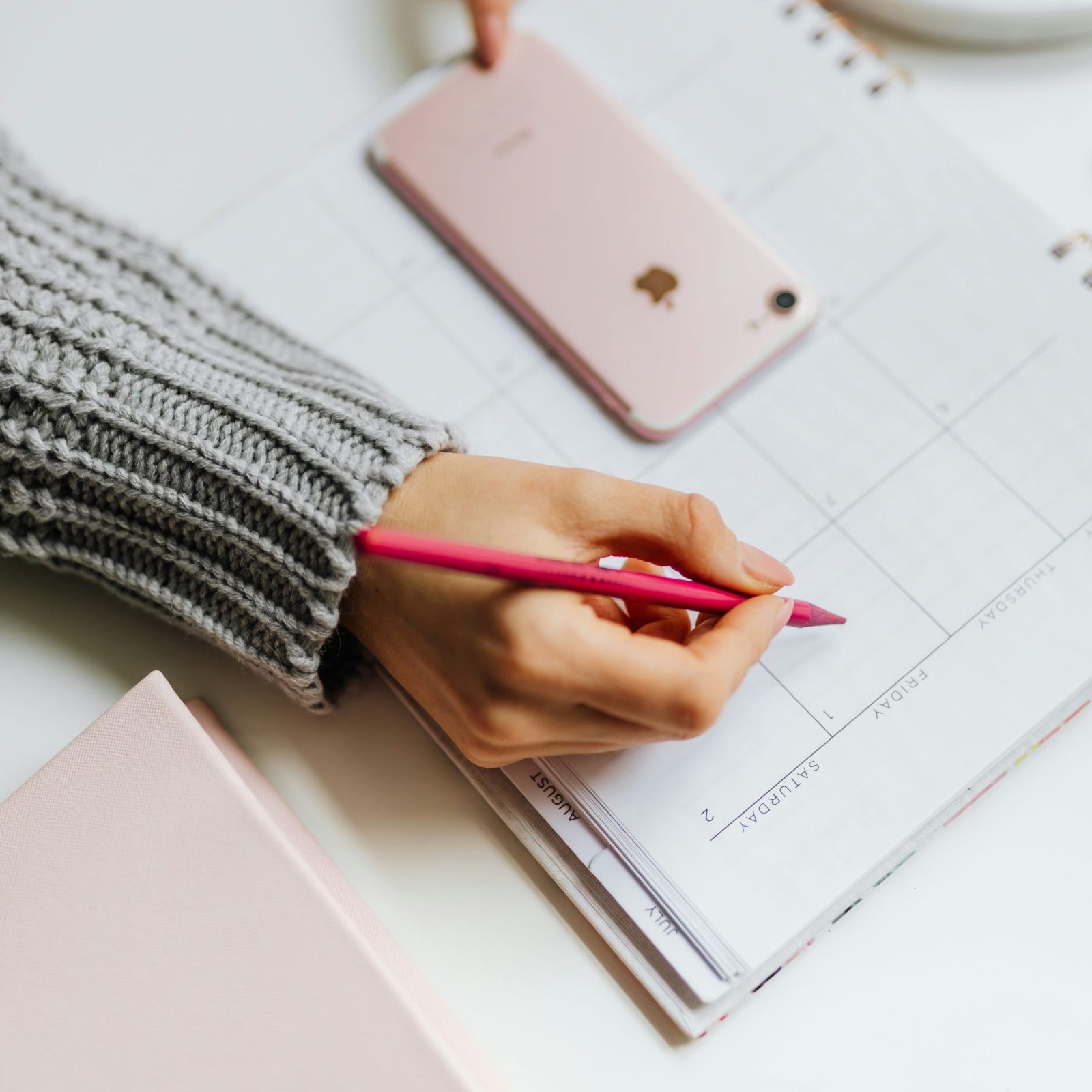 A hand holding a pink pen poised over an open calendar, with a smartphone resting nearby on the desk.