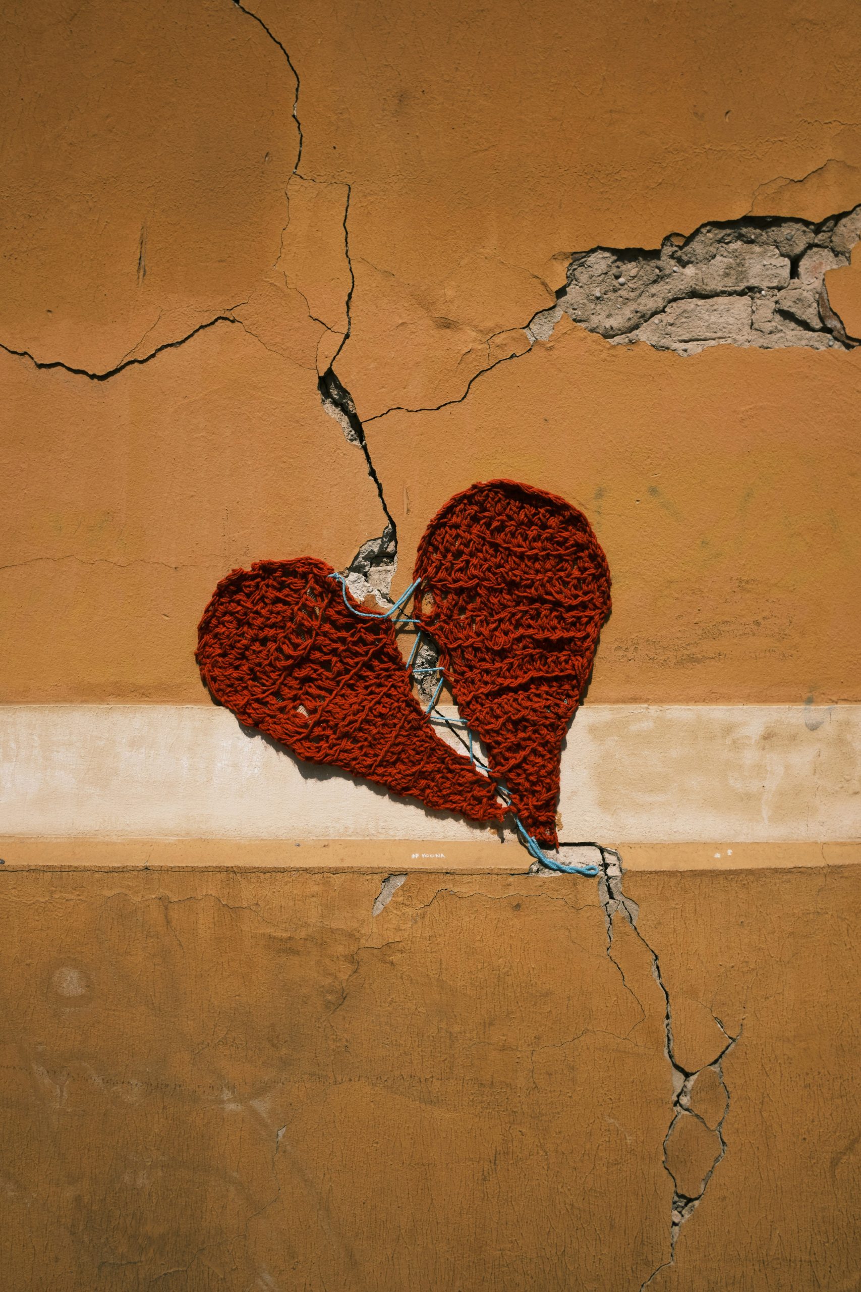 A cracked tan wall with a torn crocheted red heart stitched loosely across the fracture.