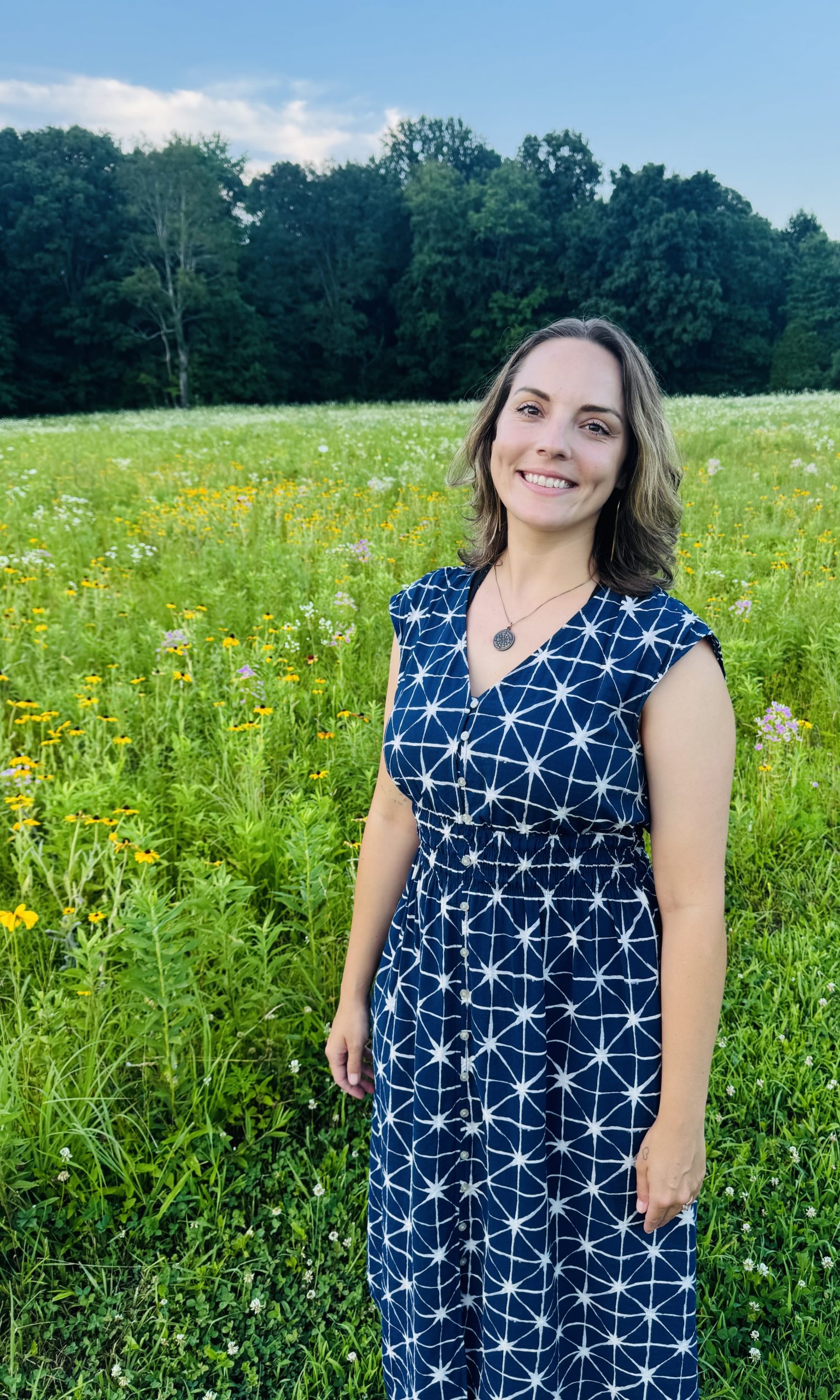 Ashlie Meredith smiling outdoors in a wildflower meadow, wearing a navy blue patterned dress.